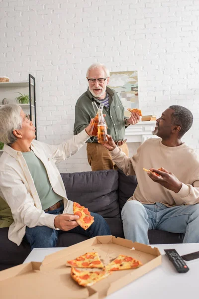 Amigos mayores multiculturales que tintinean cerveza cerca de sabrosa pizza y controlador remoto en casa — Stock Photo