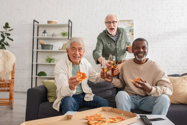 Personas mayores multiétnicas felices sosteniendo botellas de pizza y cerveza en casa - foto de stock