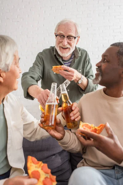 Sonriente hombre mayor tintineando cerveza con amigos interracial sosteniendo pizza borrosa en casa - foto de stock