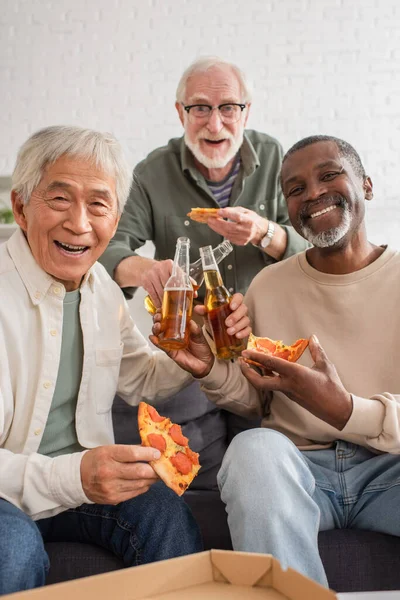 Interrazziale anziani uomini guardando la fotocamera, mentre in possesso di pizza e birra a casa — Foto stock