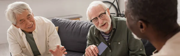 Smiling multiethnic men with playing card talking to african american friend at home, banner — Stock Photo