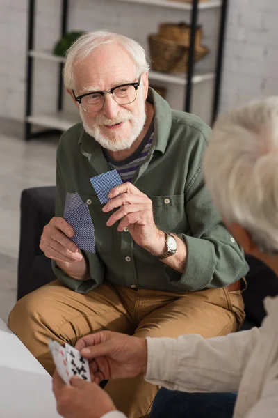 Lächelnder älterer Mann hält Spielkarten neben verschwommenem Freund zu Hause — Stockfoto