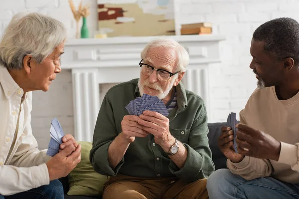 Elderly man holding playing cards near multicultural friends at home — Stock Photo