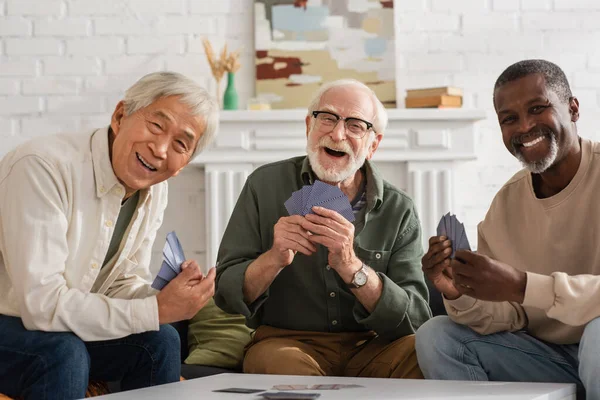 Fröhliche interrassische Freunde, die Spielkarten in der Hand halten und zu Hause in die Kamera schauen — Stockfoto