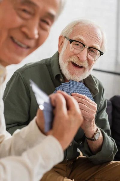 Sorrindo homem de cabelos grisalhos segurando cartas perto turvo asiático amigo em casa — Fotografia de Stock