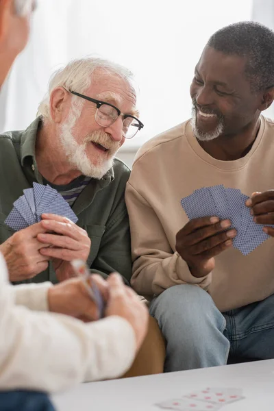 Hombres mayores multiétnicos alegres jugando a las cartas en casa — Stock Photo