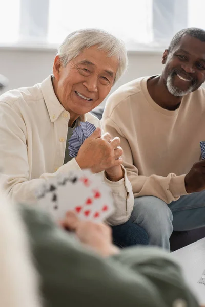 Souriant asiatique homme tenant cartes à jouer près interracial amis à la maison — Photo de stock