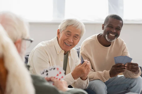 Hommes âgés multiethniques jouant aux cartes avec un ami flou à la maison — Photo de stock