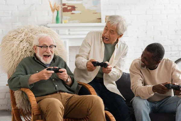 QUIIV, UCRÂNIA - OUTUBRO 12, 2021: Idosos homens idosos multiétnicos que olham para o amigo enquanto jogam videogame em casa — Fotografia de Stock