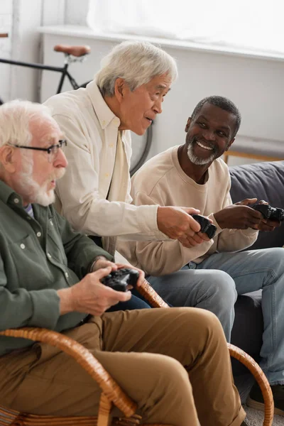 QUIIV, UCRÂNIA - OUTUBRO 12, 2021: Homem asiático focado jogando videogame com amigos idosos em casa — Fotografia de Stock