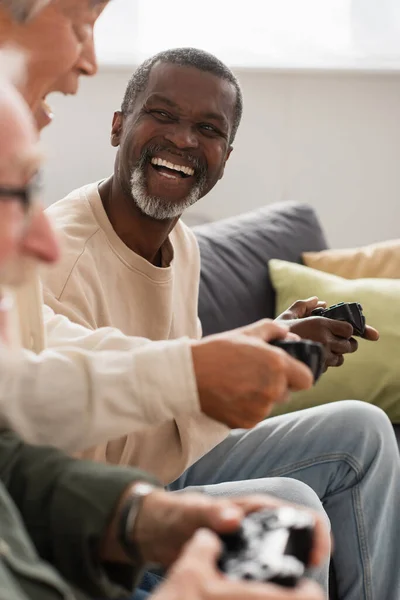 KYIV, UCRANIA - 12 de octubre de 2021: Alegre hombre afroamericano jugando videojuegos con amigos mayores interracial en casa - foto de stock