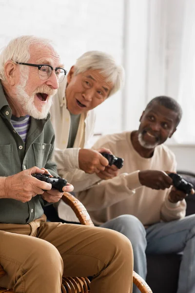 КИЕВ, УКРАИНА - 12 ОКТЯБРЯ 2021: Возбужденный мужчина играет в видеоигры со старшими межрасовыми друзьями дома — стоковое фото