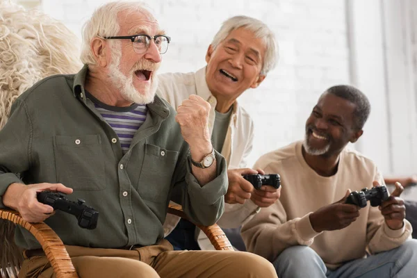 KYIV, UCRANIA - 12 DE OCTUBRE DE 2021: Emocionado hombre mayor mostrando sí gesto cerca de amigos multiétnicos con joysticks en casa - foto de stock