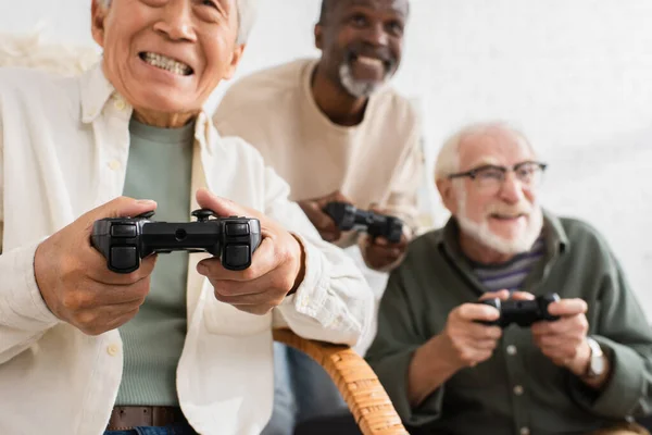 KYIV, UCRANIA - 12 de octubre de 2021: Hombre asiático mayor jugando videojuegos cerca de amigos multiétnicos borrosos en la sala de estar - foto de stock