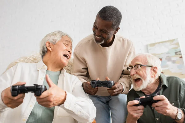 KYIV, UCRANIA - 12 de octubre de 2021: Hombre asiático positivo hablando con amigos mientras juega videojuegos en casa - foto de stock