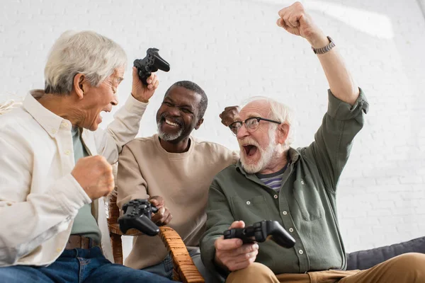 KYIV, UCRANIA - 12 DE OCTUBRE DE 2021: Hombres mayores interracial excitados sosteniendo joysticks en casa - foto de stock