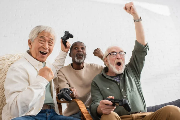 KYIV, UKRAINE - OCTOBER 12, 2021: Excited multicultural friends showing yes gesture while playing video game at home — Stock Photo