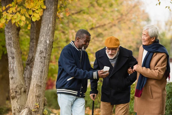 Hombre afroamericano sosteniendo teléfono inteligente cerca de amigos mayores interracial en el parque de otoño - foto de stock