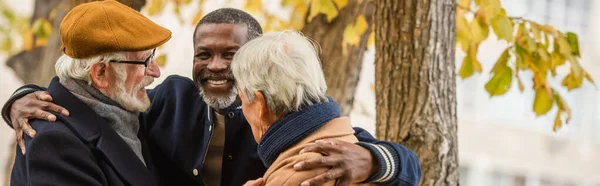 Uomo afroamericano anziano che abbraccia amici multietnici nel parco autunnale, banner — Foto stock