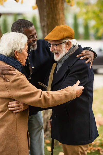 Cheerful multiethnic senior men hugging in park in autumn — Stock Photo
