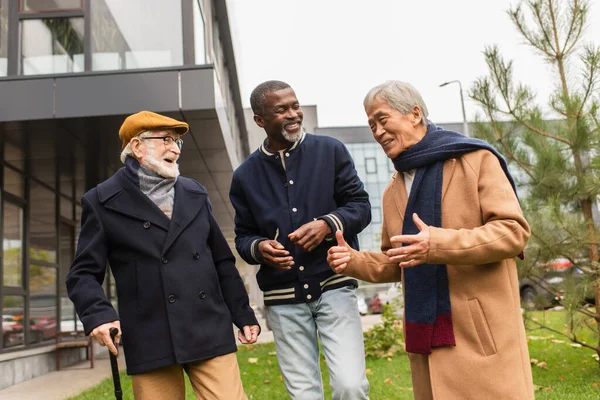 Grigio dai capelli asiatico uomo parlando con multietnico amici e mostrando come gesto su urbano strada — Foto stock