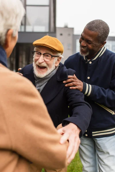 Cheerful senior man hugging blurred friend near african american man on urban street — Stock Photo