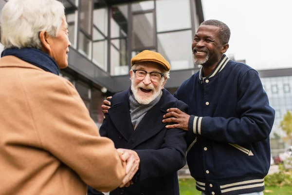 Cheerful senior man looking at camera near multiethnic friends on urban street in autumn — Stock Photo