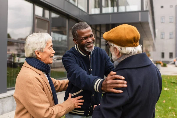 Feliz africano americano pensionista abrazando senior amigo cerca asiático hombre en urbano calle - foto de stock