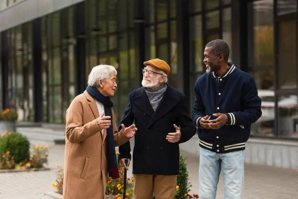 Hombres multiétnicos senior positivos hablando mientras caminan por la calle urbana - foto de stock