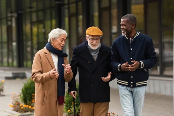 Hombre de pelo gris con bastón que habla con amigos interracial en la calle urbana - foto de stock