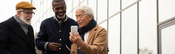 Anziano asiatico uomo utilizzando smartphone vicino interrazziale amici su urbano strada banner — Foto stock