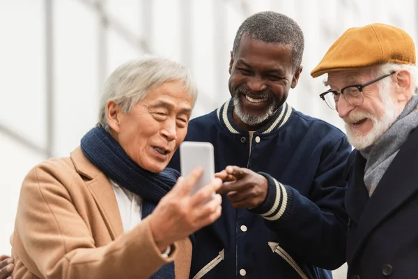 Amigos ancianos interracial positivos mirando el teléfono inteligente al aire libre - foto de stock