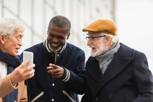 Sonriente hombre afroamericano apuntando con el dedo al teléfono inteligente cerca de amigos multiétnicos al aire libre — Stock Photo