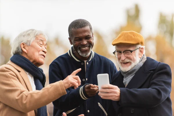 Positive interracial pensioners pointing at smartphone on urban street — Stock Photo