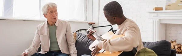Pensionistas multiétnicos tocando la guitarra acústica en el sofá en casa, pancarta - foto de stock