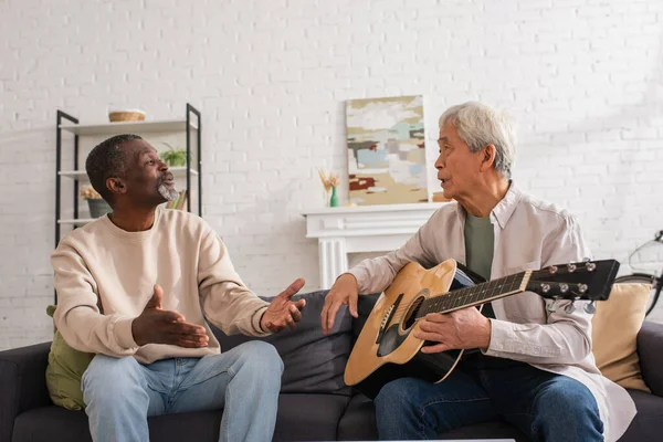 Gris peludo asiático hombre celebración acústica guitarra cerca africano americano amigo en casa - foto de stock