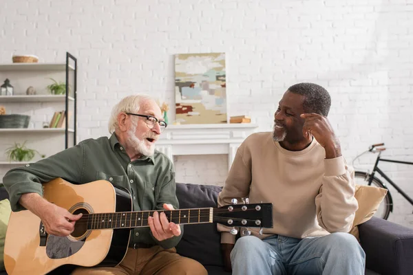 Старший мужчина играет на акустической гитаре рядом с улыбающимся африканским американским другом на диване дома — стоковое фото