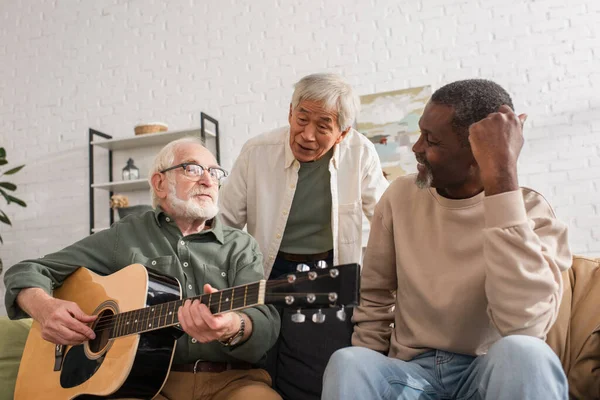 Pensionistas Interraciales mirando a un amigo tocando la guitarra acústica en casa - foto de stock