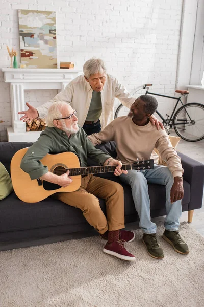 Asian man singing near multiethnic friends with acoustic guitar on couch at home — Stock Photo