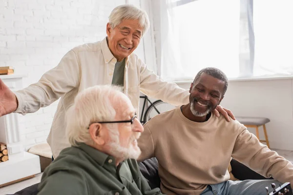Positive interracial pensioners looking at blurred friend near acoustic guitar at home — Stock Photo