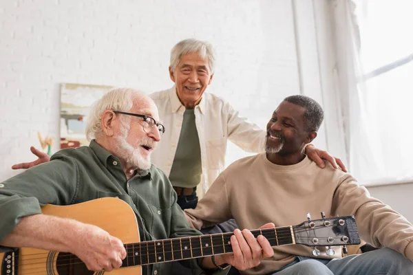 Hombre de pelo gris tocando la guitarra acústica cerca de amigos interracial en casa — Stock Photo