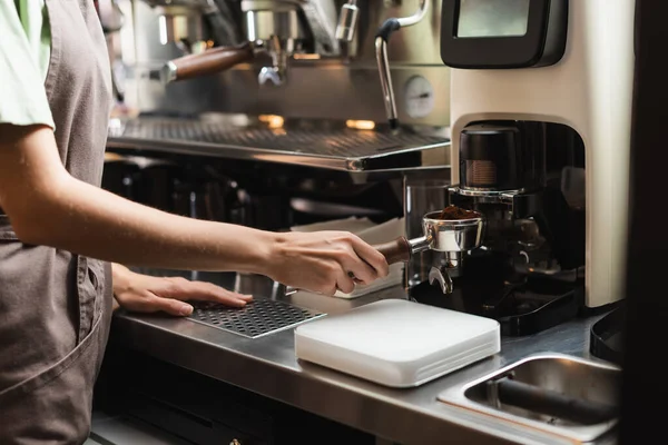 Cropped view of barista in apron holding portafilter near coffee grinder in cafe — Stock Photo