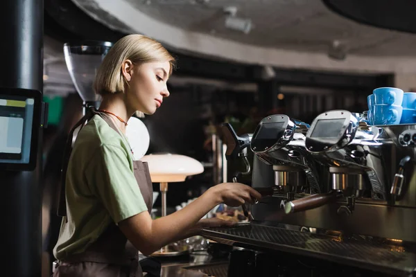 Vista lateral de un joven camarero trabajando con una cafetera en la cafetería - foto de stock