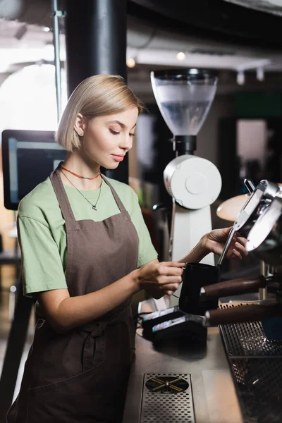 Jovem barista em avental segurando varinha de vapor de máquina de café e jarro de leite no café — Fotografia de Stock