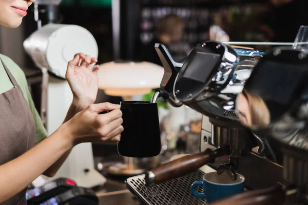 Cropped view of young barista holding jug near steam wand of coffee machine in cafe — Stock Photo