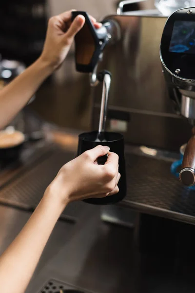 Cropped view of barista holding jug near blurred steam wand of coffee machine in cafe — Stock Photo