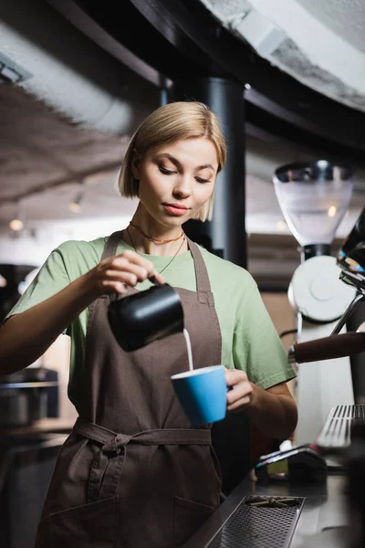 Barista bionda in grembiule versando latte in tazza vicino alla macchina del caffè in caffè — Foto stock