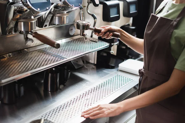 Cropped view of barista in apron holding portafilter near coffee machine in cafe — Stock Photo
