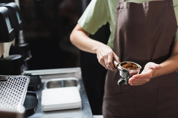 Ausgeschnittene Ansicht eines verschwommenen Barista, der einen Filter mit Kaffee in der Nähe der Kaffeemaschine in der Cafeteria hält — Stockfoto