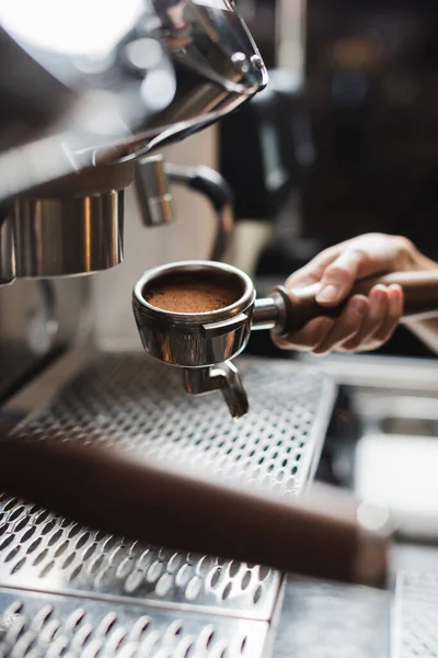 Cropped view of barista holding portafilter with coffee near blurred coffee machine on foreground in cafe — Stock Photo